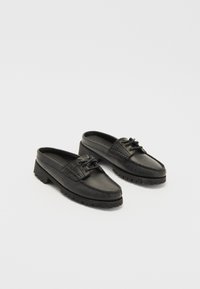 Chaussons en cuir noir à enfilage avec un design de style mocassin, présentant des coutures décoratives, un devant à lacets et des semelles en caoutchouc texturé.