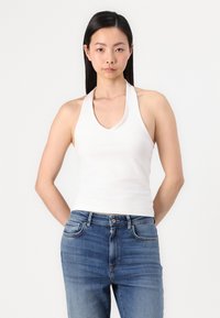 Bijela halter-top majica s V-izrezom, uparen s trapericama srednjeg struka u plavoj boji, koje sadrže suptilno izbljeđivanje i klasičan dizajn s pet džepova.