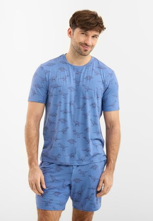 Ung mand iført blå matchende shorts og T-shirt med mørke dinosaurmønstre, smilende, stående mod en hvid baggrund.