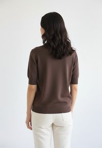 Kobieta o falujących ciemnych włosach, ubrana w brązowy sweter z krótkim rękawem i białe spodnie, stojąca tyłem na jasnym, jednolitym tle.