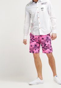 Vit långärmad skjorta med färgglada grafiska detaljer och krage, ihop med rosa shorts med tropiskt svart blommönster och vita sneakers.