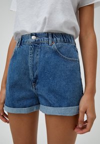 Pantalones cortos de mezclilla en azul medio, con cintura alta y puños doblados. Cierre de botón y bolsillos frontales. Tela de algodón con ligera textura.