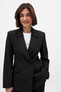 Melnā žakete, kas izgatavota no teksturēta auduma, ar divu pogu aizdari, lielām atlokām un divām priekšējām kabatām. Valkāta virs baltas krekla.