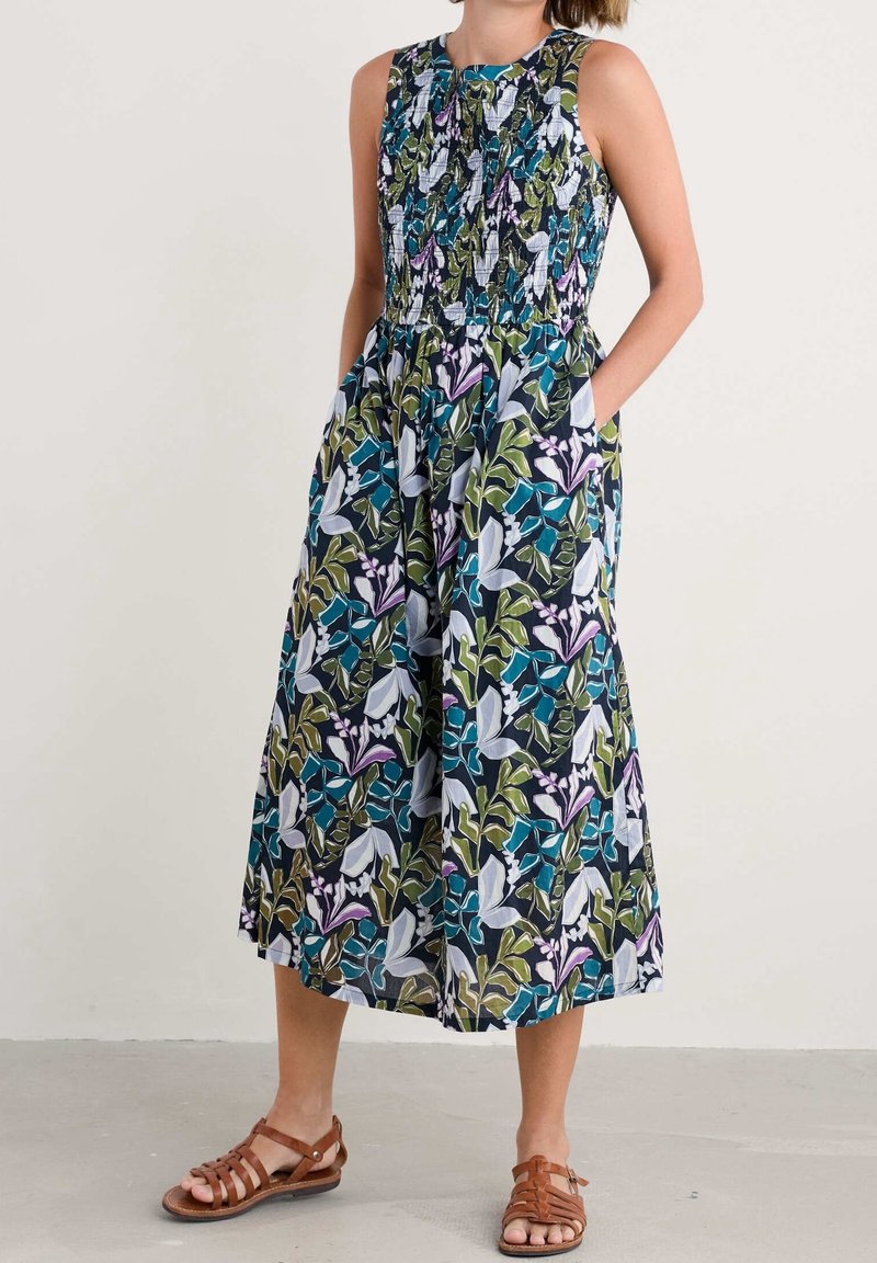 Robe midi sans manches avec un corsage smocké et un imprimé de feuilles tropicales vertes, bleues, blanches et violettes, portée avec des sandales marron avec lanières.