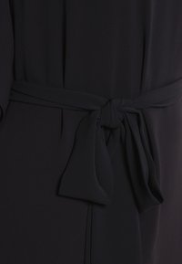 Robe en tissu foncé avec un lien à la taille. Texture lisse, design fluide et détail de nœud simple. Style minimaliste, sans motifs ni éléments métalliques visibles.