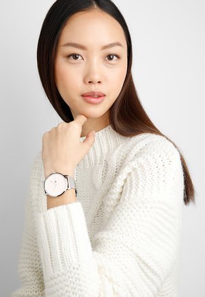 Montre argentée ronde avec un cadran blanc et un design minimaliste, dotée d'un bracelet en maille. Le sujet porte un pull en laine blanc.