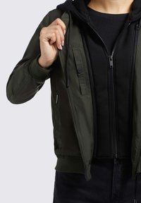 Maslinasto zelena bomber jakna s patentnim zatvaračem, rebrastim manžetama i prednjim džepom, složena preko crnog dukserice s patentnim zatvaračem.