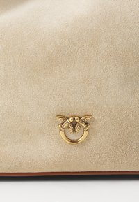 Borsa a mano in suede beige con un design circolare in metallo dorato e due motivi di uccelli. Texture liscia e morbida con un'eleganza sobria.
