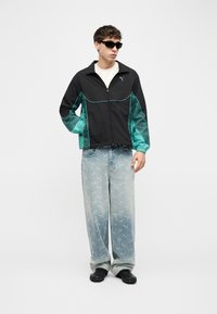 Jeune homme portant une veste zippée noire et bleu sarcelle, un jean large bleu clair avec un imprimé logo sur l'ensemble, des chaussures noires et des lunettes de soleil noires, debout devant un fond blanc.