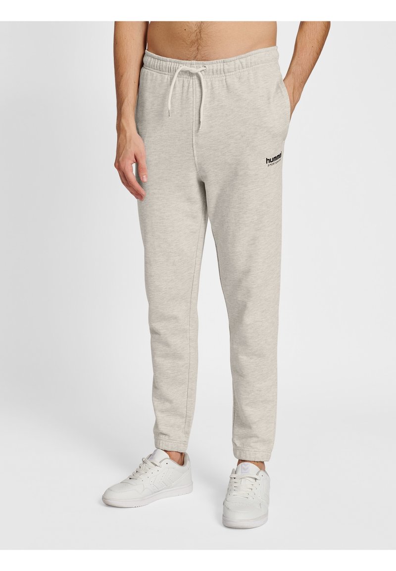 Homme portant un pantalon de jogging gris clair avec une taille à cordon et des baskets blanches, debout les mains partiellement dans les poches sur un fond uni.