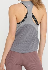 Top sportivo grigio con schiena perforata, design racerback e spalline con logo Nike nero, abbinato a leggings lisci di un rosa chiaro.