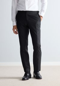 Pantaloni neri su misura con una texture liscia, dotati di un davanti piatto e tasche angolate. Abbinati a scarpe in pelle nera lucida.