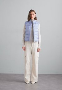 Hellblauer Steppweste über einem cremefarbenen Pullover und strukturierten weißen, weit geschnittenen Hosen mit seitlichen Taschen. Als Schuhe werden spitze beige Schuhe getragen.
