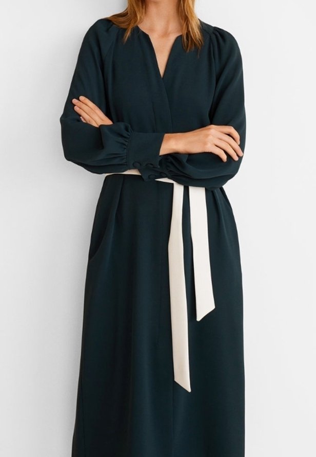 Femme portant une longue robe noire à manches bouffantes et une ceinture blanche nouée à la taille, les bras croisés devant elle.