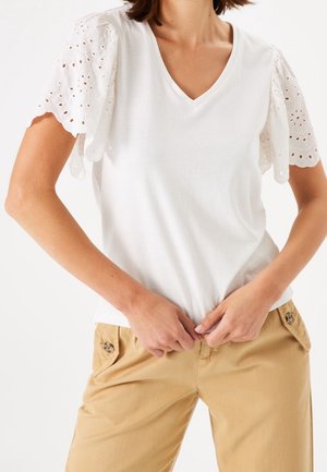 Biała t-shirt z dekoltem w serek i haftowanymi rękawami z ażurowymi wstawkami, w połączeniu z beżowymi spodniami z kieszeniami na guziki. Gładka tkanina, luźny krój.