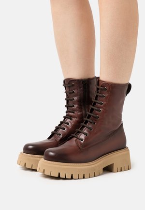 Nohy v tmavě hnědých kožených šněrovacích botách s tlustými béžovými masivními podrážkami a bočními zipy na jednobarevném pozadí.