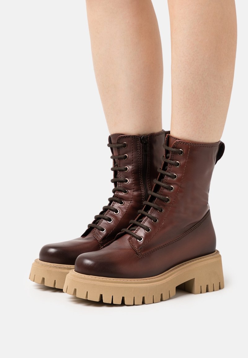 Jambes portant des bottes en cuir marron foncé à lacets, avec des semelles épaisses beiges et des fermetures éclair latérales, sur un fond uni.