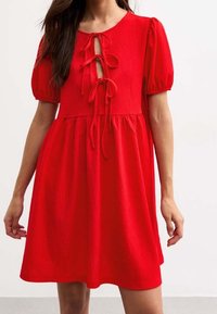 Robe courte rouge avec encolure ronde, manches bouffantes et détail à nouer à l'avant. Tissu texturé avec une jupe évasée de longueur genou.