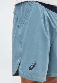 Κοντινή λήψη αθλητικού σορτς σε ανοιχτό γαλάζιο χρώμα με πλευρική τσέπη και μαύρο λογότυπο Asics στο αριστερό πόδι.