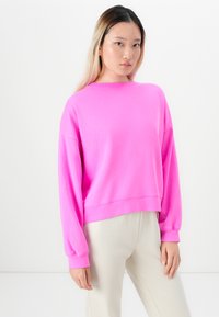 Young woman with long hair wearing a bright pink long-sleeve sweatshirt and light beige pants against a white background.