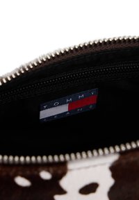 Interiorul unui săculeț din piele de vacă maro și alb, prevăzut cu un fermoar negru și o etichetă „TOMMY JEANS” cu accente albastre, roșii și albe.