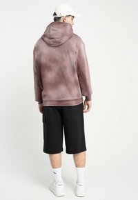 Przesadnie duża, wyblakła mauve bluza z kapturem, połączona z czarnymi szortami cargo. Model ma na sobie białe trampki i skarpetki sportowe. Minimalistyczny design.