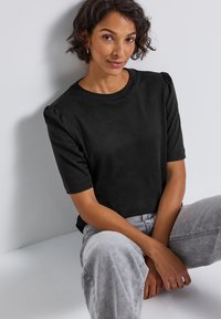 Sort kortærmet top med rund halsudskæring, der har subtile puffede ærmedetaljer. Bæres med lysegrå jeans, og stoffet fremstår glat.