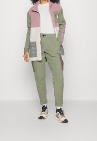 Fleece jacket in pink, green, and white with a collar and pockets; paired with green fleece pants featuring cargo pockets. Neutral sneakers worn.