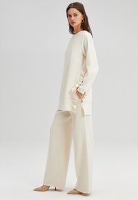 Cream-colored, long-sleeve top with ruffled side accents; paired with wide-leg, matching pants and pointed beige heels. Smooth texture.