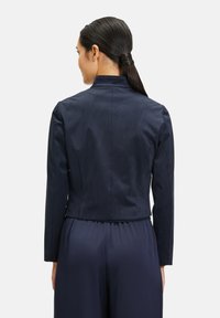 Marinblå kort jacka med långa ärmar och krage. Har en slät, matt yta. Bakåtsidan visar skräddarsydd sömnad och rena linjer.