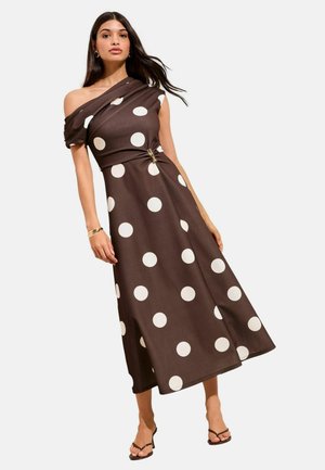 Donna che indossa un abito monospalla marrone con grandi pois bianchi, una fibbia in vita e tacchi aperti neri.