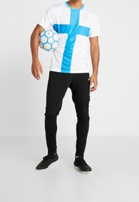 Valkoinen jalkapallopaita sinisellä ristillä, logo rinnan kohdalla, mustat kapenevat housut ja mustat nappulakengät. Pelaaja pitää sinistä jalkapalloa.