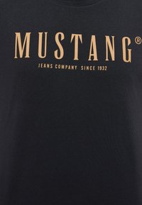 Zwarte T-shirt met een groot, gouden "MUSTANG" logo en kleinere tekst "JEANS COMPANY SINCE 1932." Soepele katoentextuur.