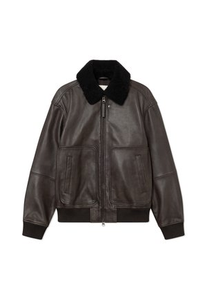 Jeune homme portant une veste en cuir noire avec col en fourrure, une chemise rose à carreaux, un pantalon noir à larges jambes à fines rayures, une casquette noire et des chaussures.