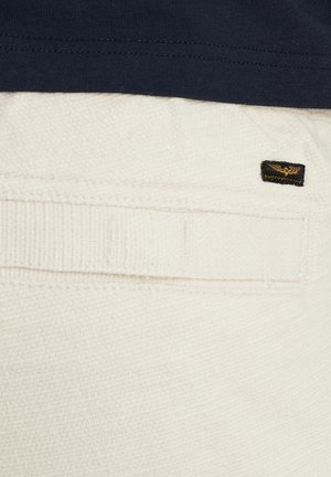 White textured fabric, featuring a visible back pocket with a stitched detail and a black logo label in the corner. Navy top partially visible.