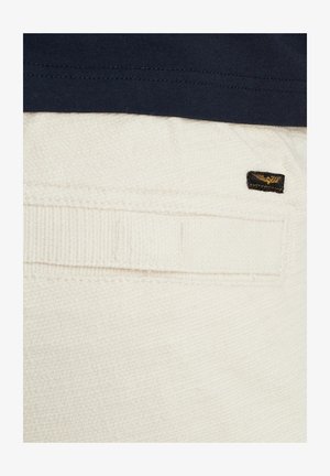 Tessuto bianco testurizzato, con una tasca posteriore visibile con dettagli cuciti e un'etichetta logo nera all'angolo. Parte di un top blu scuro è parzialmente visibile.