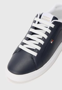 Tamsiai mėlynas odinis sportinis batelis su baltais raišteliais, balta padu ir Tommy Hilfiger logotipu ant liežuvėlio ir šono.