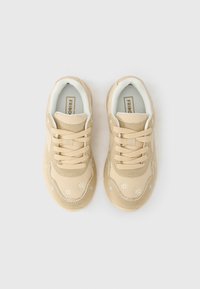 Paire de baskets beige avec lacets à l'avant et petite broderie de fleurs blanches sur le dessus, vues de dessus sur un fond blanc.