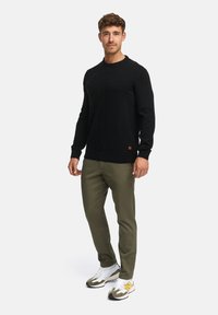 Un pull en tricot noir avec un col rond, un pantalon vert olive et des baskets blanches avec des accents jaunes, comportant une petite étiquette de marque sur le pull.