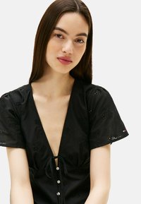 Zwarte jurk met korte mouwen en een diepe V-hals, met een strik in de taille en decoratieve bloemenborduring. Knopen sluiting aan de voorkant.