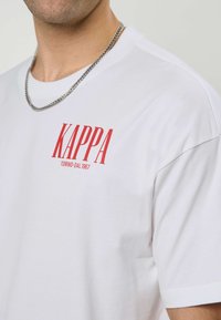 Άνδρας που φοράει λευκό μπλουζάκι Kappa με κόκκινο λογότυπο και κείμενο "KAPPA TORINO DAL 1967" και ασημένιο αλυσιδωτό κολιέ σε ανοιχτό φόντο.