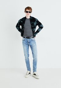Black and green tie-dye jacket, fitted gray t-shirt, distressed light blue jeans, and cream sneakers with black accents. Accessories include sunglasses.