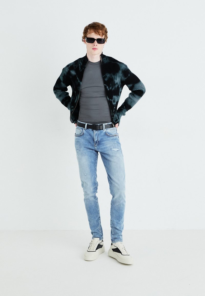 Veste tie-dye noire et verte, t-shirt gris ajusté, jeans bleu clair déchirés et baskets crème avec accents noirs. Les accessoires incluent des lunettes de soleil.