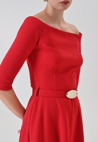 Rotes Kleid mit schulterfreiem Ausschnitt und 3/4-Ärmeln. Es hat eine betonte Taille mit einer metallischen Gürtelschnalle. Glatte Stoffstruktur.