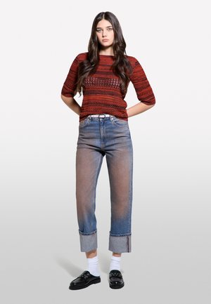 Femme aux longs cheveux foncés porte un haut rouge à manches courtes à motifs, un jean bleu avec revers, des chaussettes blanches et des chaussures noires faciles à enfiler sur un fond uni.