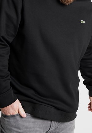 Homme portant un sweat Lacoste noir avec un logo crocodile vert et un jean gris, main dans la poche, barbe taillée visible.