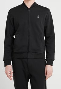 Svart zip-up jacka med sidofickor och vitt broderat polo-spelare logotyp på vänster bröst, bärs med svarta byxor.