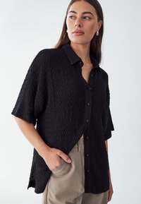 Camisa negra texturizada de manga corta y cierre de botones en la parte delantera. Presenta un cuello con solapa y un corte holgado, ideal para combinar con pantalones de color claro.