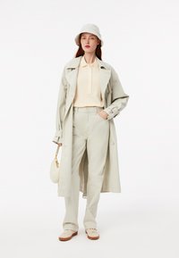Lichtgrijze trenchcoat, lichtbeige polo, bijpassende hoog-waist broek en crèmekleurige sneakers. Houdt een kleine ronde tas vast; een bucket hat maakt de outfit compleet.