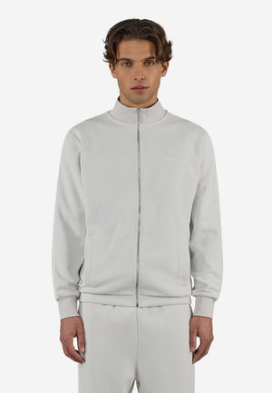 Mann trägt hellgraue Reißverschlussjacke und passende Hose, steht frontal vor einem schlichten weißen Hintergrund.